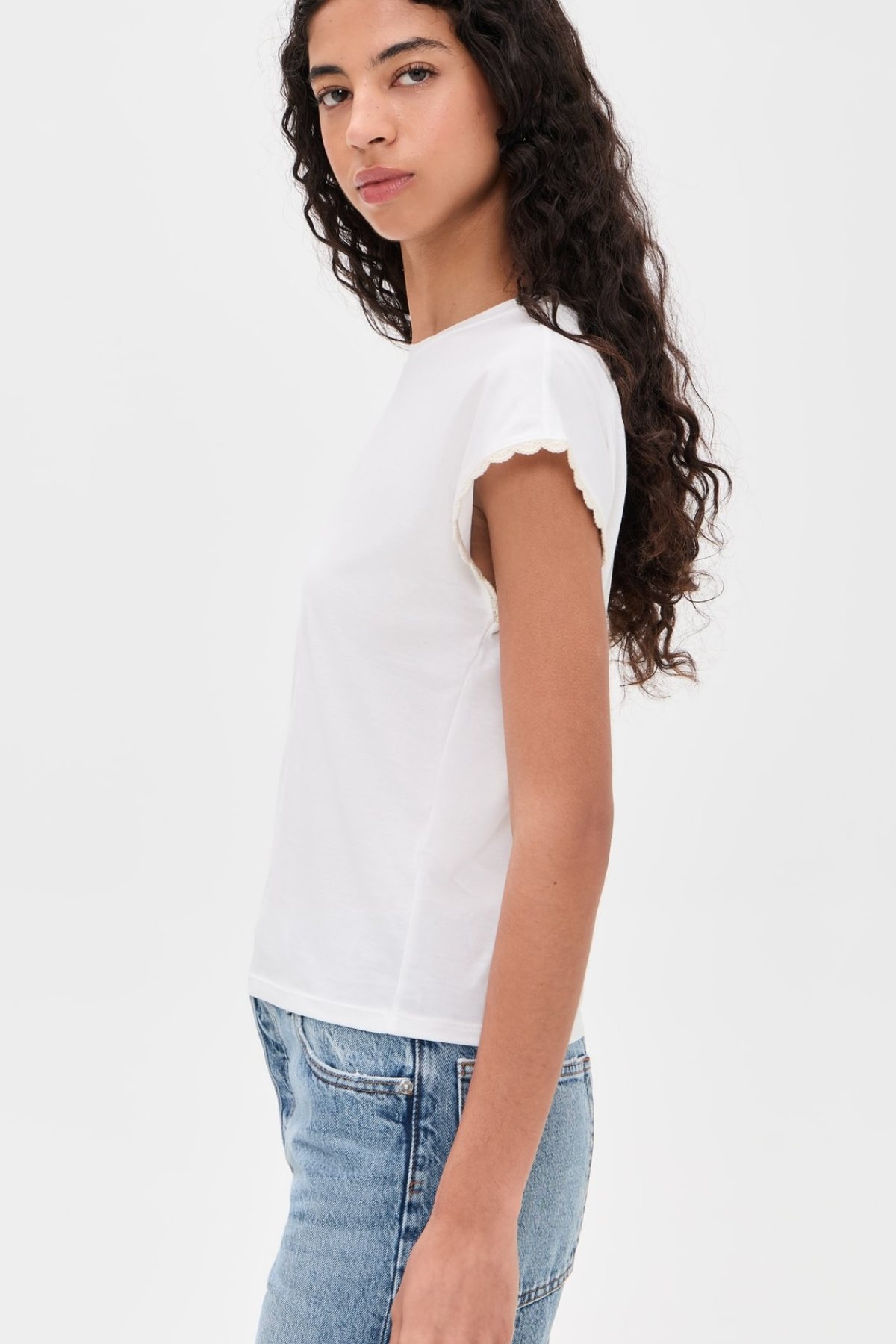 A woman wearing a white short-sleeve t-shirt with delicate scalloped edges on the sleeves, paired with light blue jeans, shown from a side profile against a plain white background.