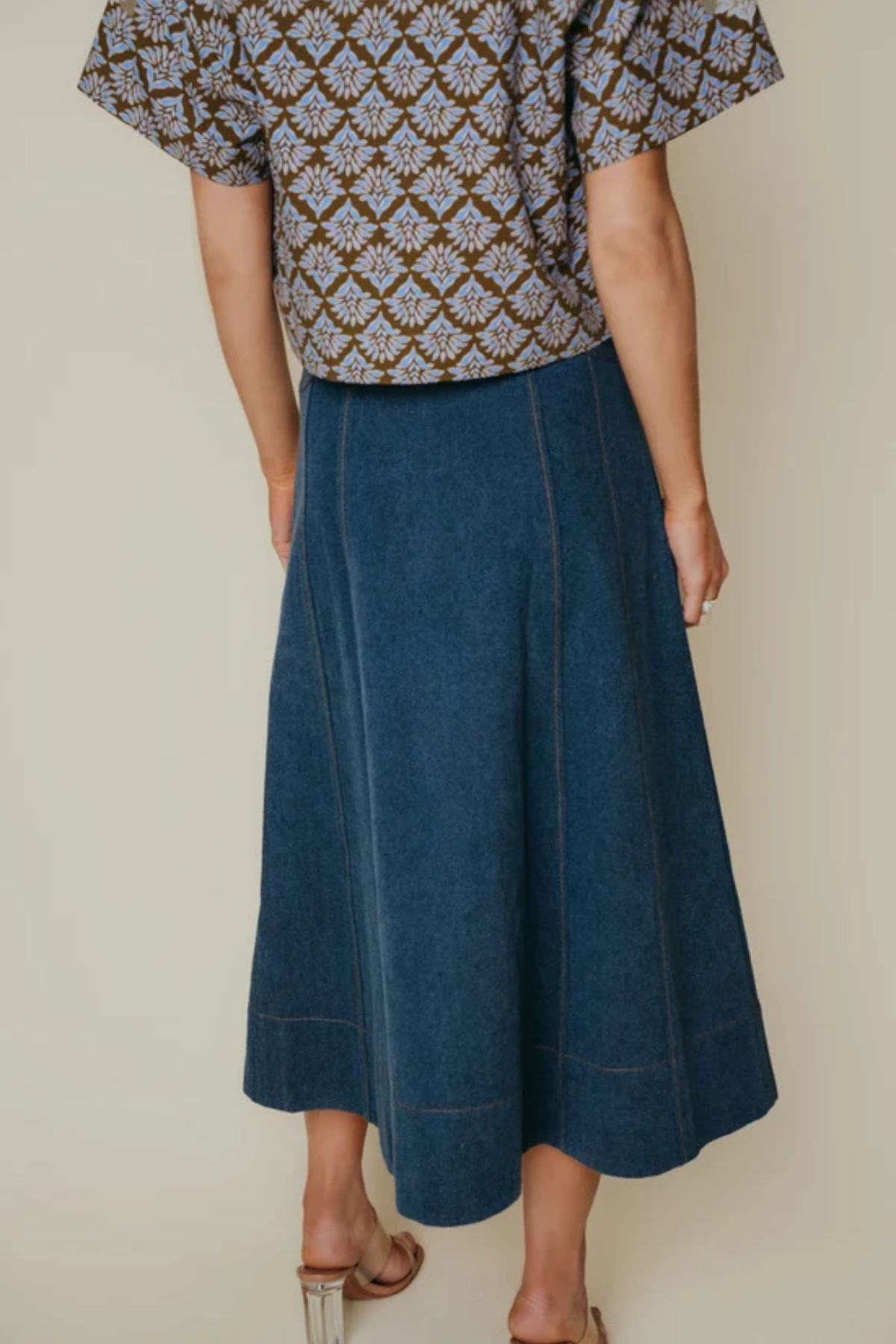 A person shown from the back wearing a dark blue denim midi skirt with a patterned top, standing against a plain beige studio background.