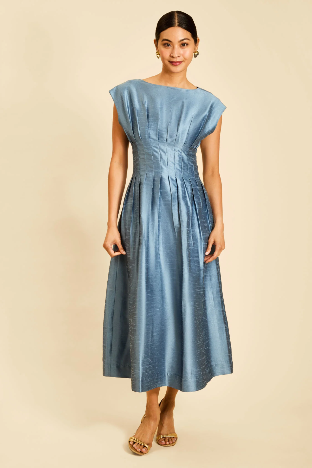 A woman modeling a light blue midi dress with a gathered waist and cap sleeves, standing against a solid beige background.