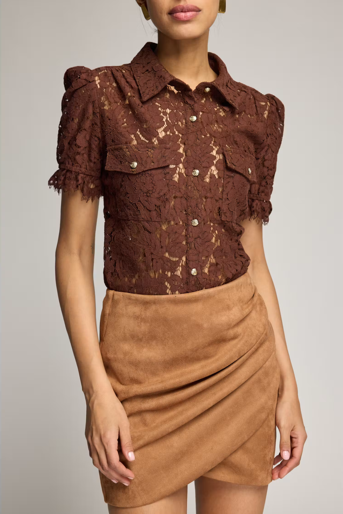 A model wearing a brown lace short-sleeve button-up blouse with ruffled sleeves and a tan faux-suede wrap mini skirt, photographed against a light gray studio background.