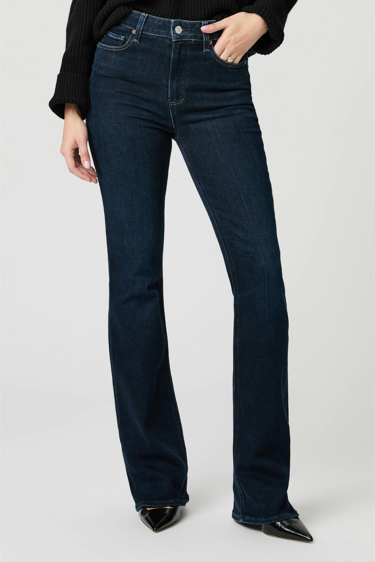 A model wearing dark blue flare jeans, a black long-sleeve top, and black pointed-toe heels, standing against a light gray studio background.