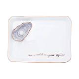 A white rectangular ceramic tray with rounded corners, featuring a raised 3D oyster shell design in the top-left corner and the phrase 'the world is your oyster' in rose gold script along the bottom edge.