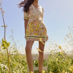 Woman in a patterned dress standing on a chair in a field with a clear blue sky.