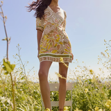 Woman in a patterned dress standing on a chair in a field with a clear blue sky.