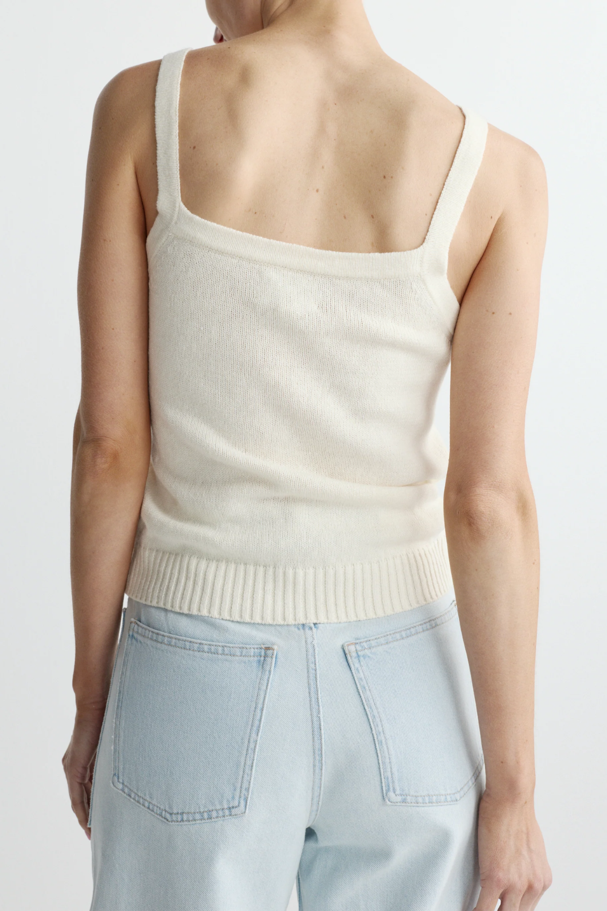 Person wearing a cream-colored knit tank top and light blue jeans on a white background