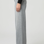 A side profile view of a model wearing light gray wide-leg denim jeans, a black long-sleeve top, and black pointed-toe heels against a light gray studio background.