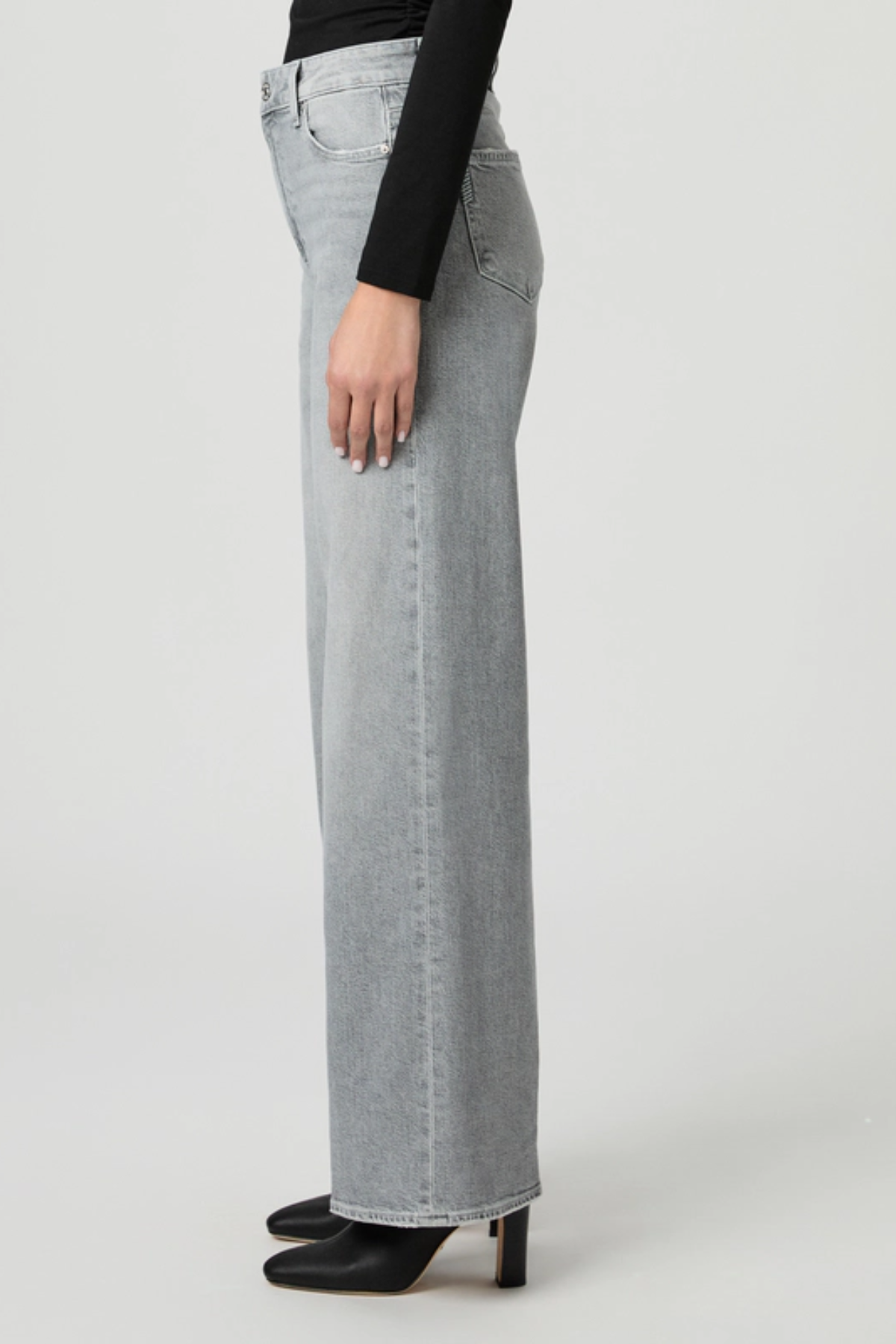 A side profile view of a model wearing light gray wide-leg denim jeans, a black long-sleeve top, and black pointed-toe heels against a light gray studio background.