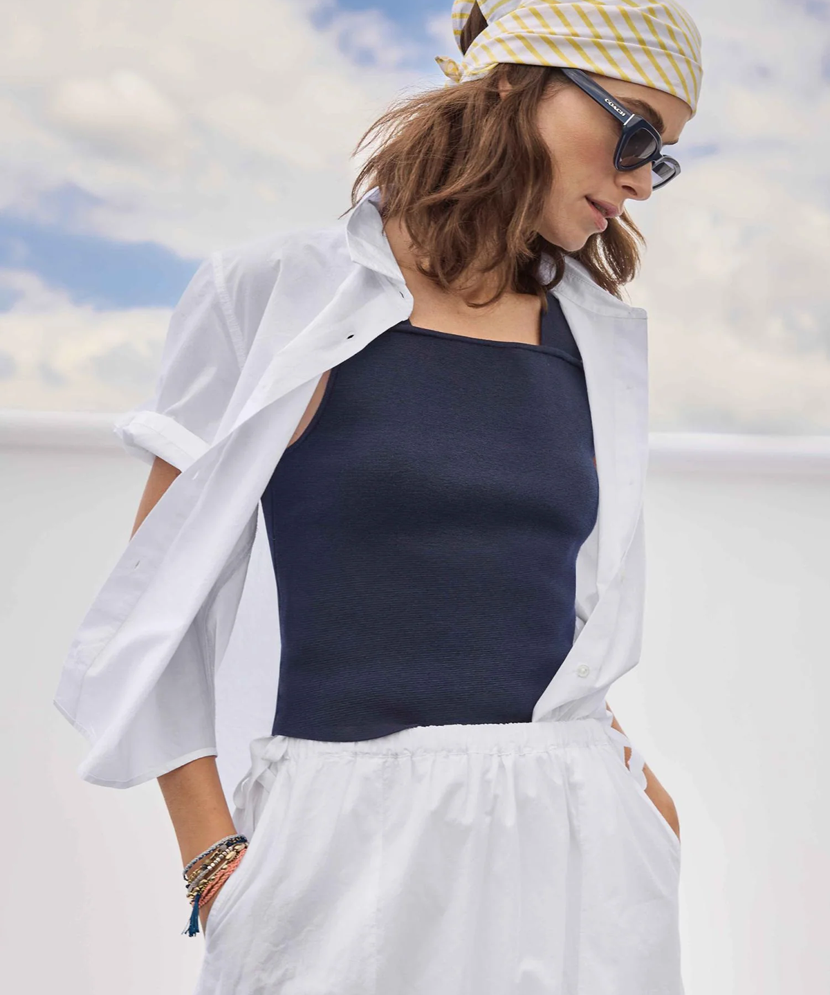 Woman wearing a white shirt, navy tank top, and white pants with a yellow and white patterned headscarf against a blue sky.
