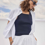 Woman wearing a white shirt, navy tank top, and white pants with a yellow and white patterned headscarf against a blue sky.