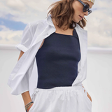 Woman wearing a white shirt, navy tank top, and white pants with a yellow and white patterned headscarf against a blue sky.