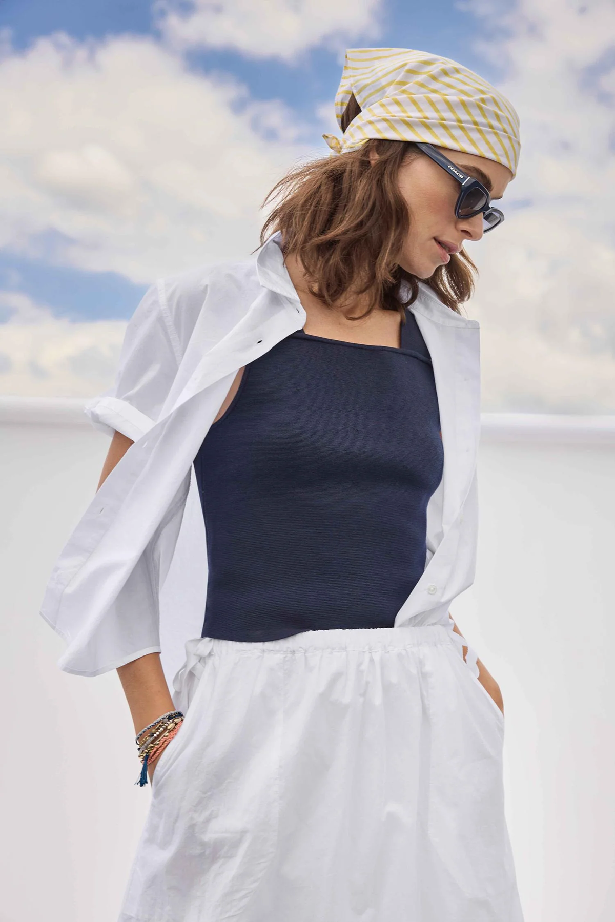 Woman wearing a white shirt, navy tank top, and white pants with a yellow and white patterned headscarf against a blue sky.