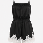 A black satin-like romper with thin shoulder straps, a gathered elastic waist, and white floral lace trim along the hem, shown on a plain light background.