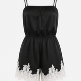 A black satin-like romper with thin shoulder straps, a gathered elastic waist, and white floral lace trim along the hem, shown on a plain light background.