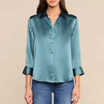 A woman modeling a teal satin button-down shirt with rolled cuffs, paired with blue jeans, against a solid beige background.