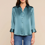 A woman modeling a teal satin button-down shirt with rolled cuffs, paired with blue jeans, against a solid beige background.