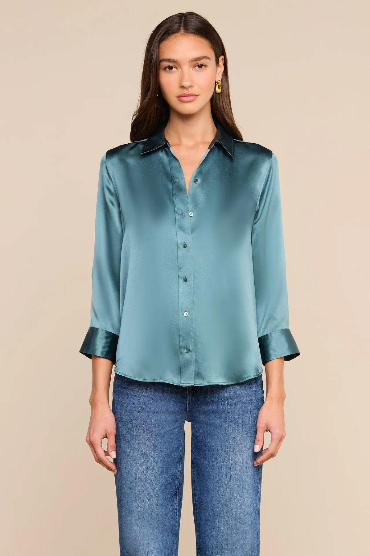 A woman modeling a teal satin button-down shirt with rolled cuffs, paired with blue jeans, against a solid beige background.
