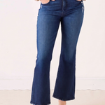 A person wearing dark blue flare jeans with a tan sweater and brown pointed flats, standing against a plain off-white wall.