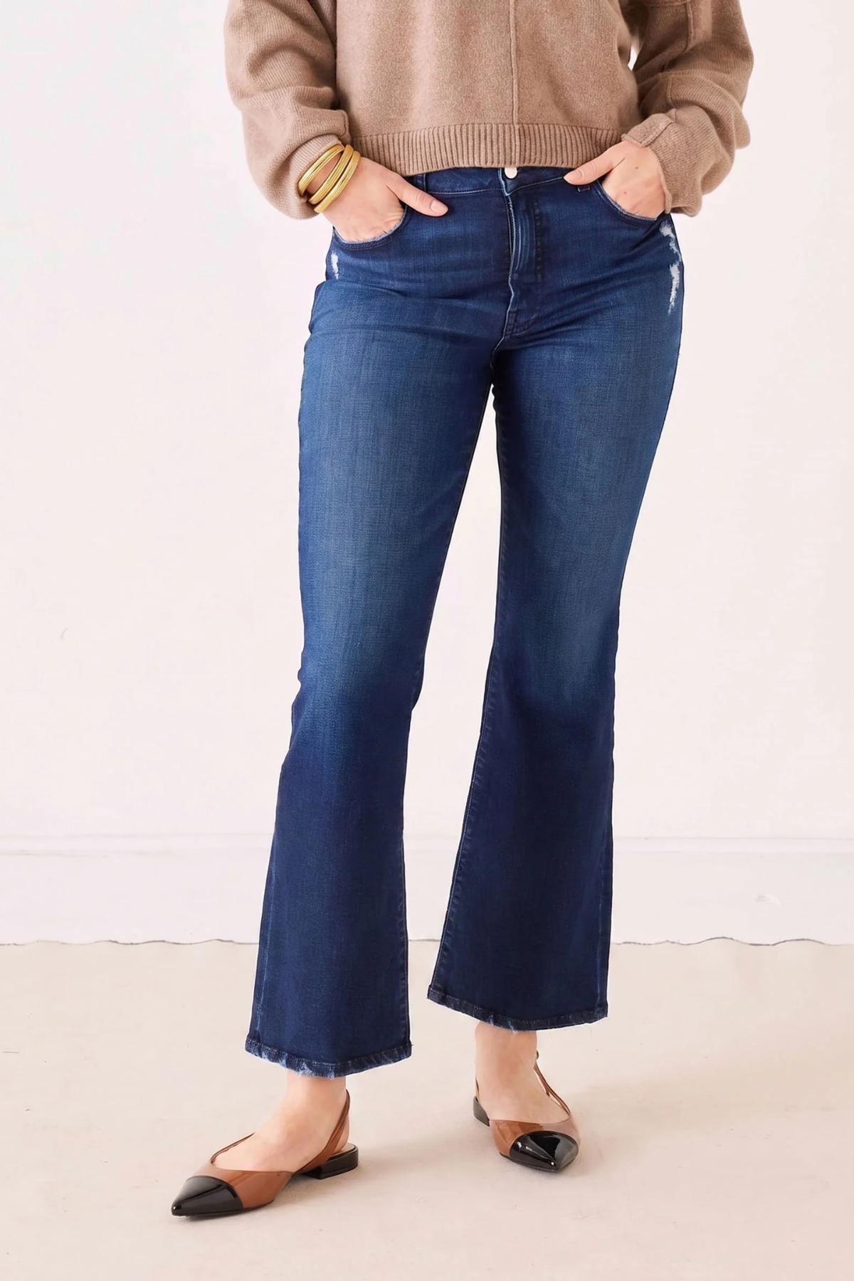 A person wearing dark blue flare jeans with a tan sweater and brown pointed flats, standing against a plain off-white wall.
