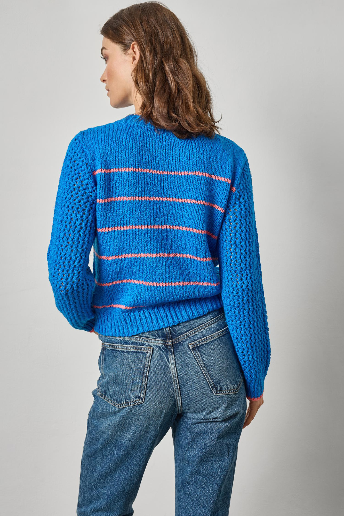 Person wearing a blue sweater with pink stripes and jeans on a gray background