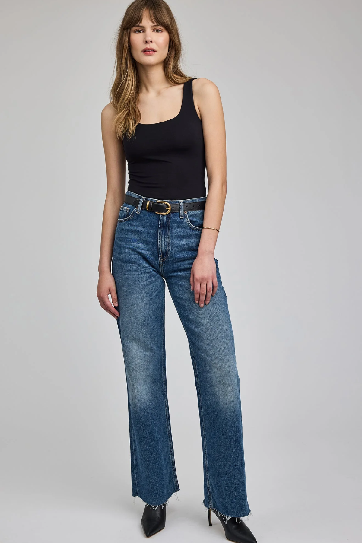 A woman modeling a pair of medium-wash blue wide-leg jeans with a black tank top and black pointed-toe heels, standing against a light gray studio background.