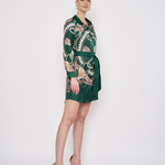 A woman modeling a dark green wrap-style mini dress with a beige and white abstract pattern, paired with beige high-heeled sandals, against a plain white studio background.