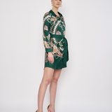 A woman modeling a dark green wrap-style mini dress with a beige and white abstract pattern, paired with beige high-heeled sandals, against a plain white studio background.