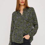 A woman modeling a long-sleeve button-up blouse with an olive green base and a black and white floral pattern, paired with dark gray jeans, against a plain light gray studio background.