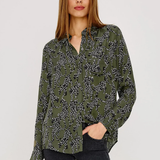 A woman modeling a long-sleeve button-up blouse with an olive green base and a black and white floral pattern, paired with dark gray jeans, against a plain light gray studio background.