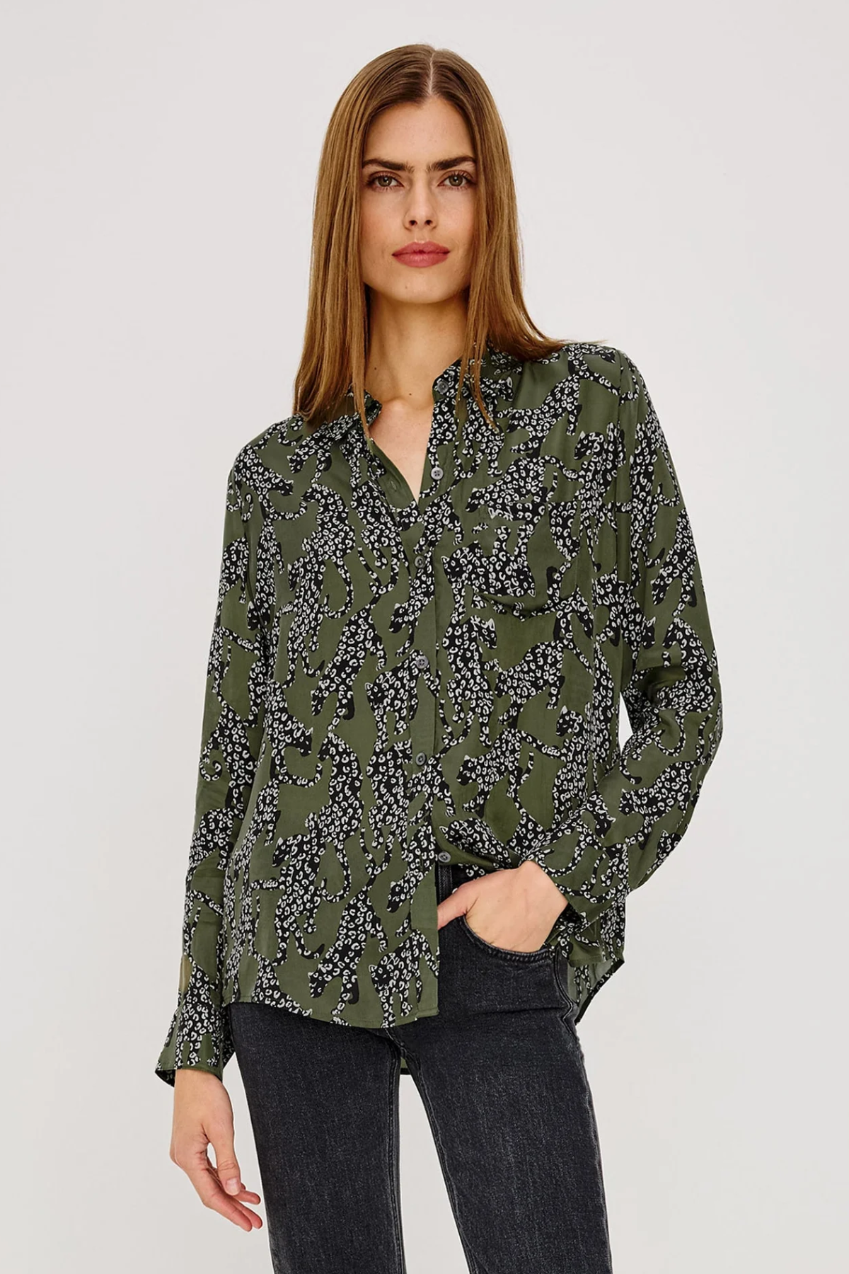 A woman modeling a long-sleeve button-up blouse with an olive green base and a black and white floral pattern, paired with dark gray jeans, against a plain light gray studio background.