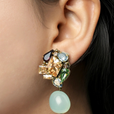 A close-up of a model's ear wearing a statement dangle earring with multiple colorful gemstones and a light green bead drop.
