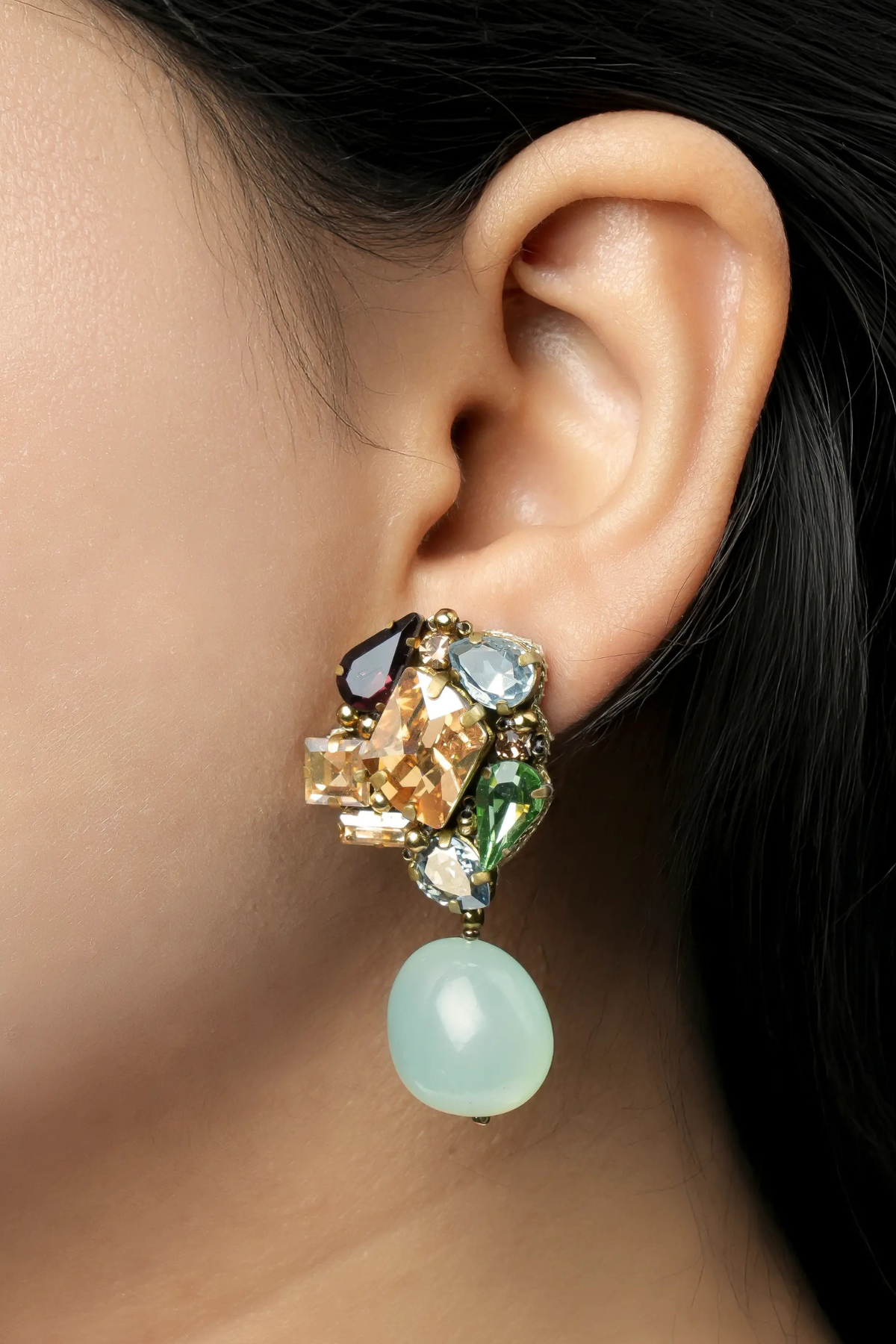 A close-up of a model's ear wearing a statement dangle earring with multiple colorful gemstones and a light green bead drop.
