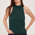 A woman modeling a sleeveless, high-neck cable-knit sweater vest in dark forest green, paired with dark blue jeans, against a plain light gray studio background.