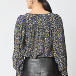 A woman shown from the back wearing a long-sleeve blouse with a dense multicolor floral print on a dark background, paired with a black leather skirt, against a light gray studio background.
