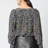A woman shown from the back wearing a long-sleeve blouse with a dense multicolor floral print on a dark background, paired with a black leather skirt, against a light gray studio background.