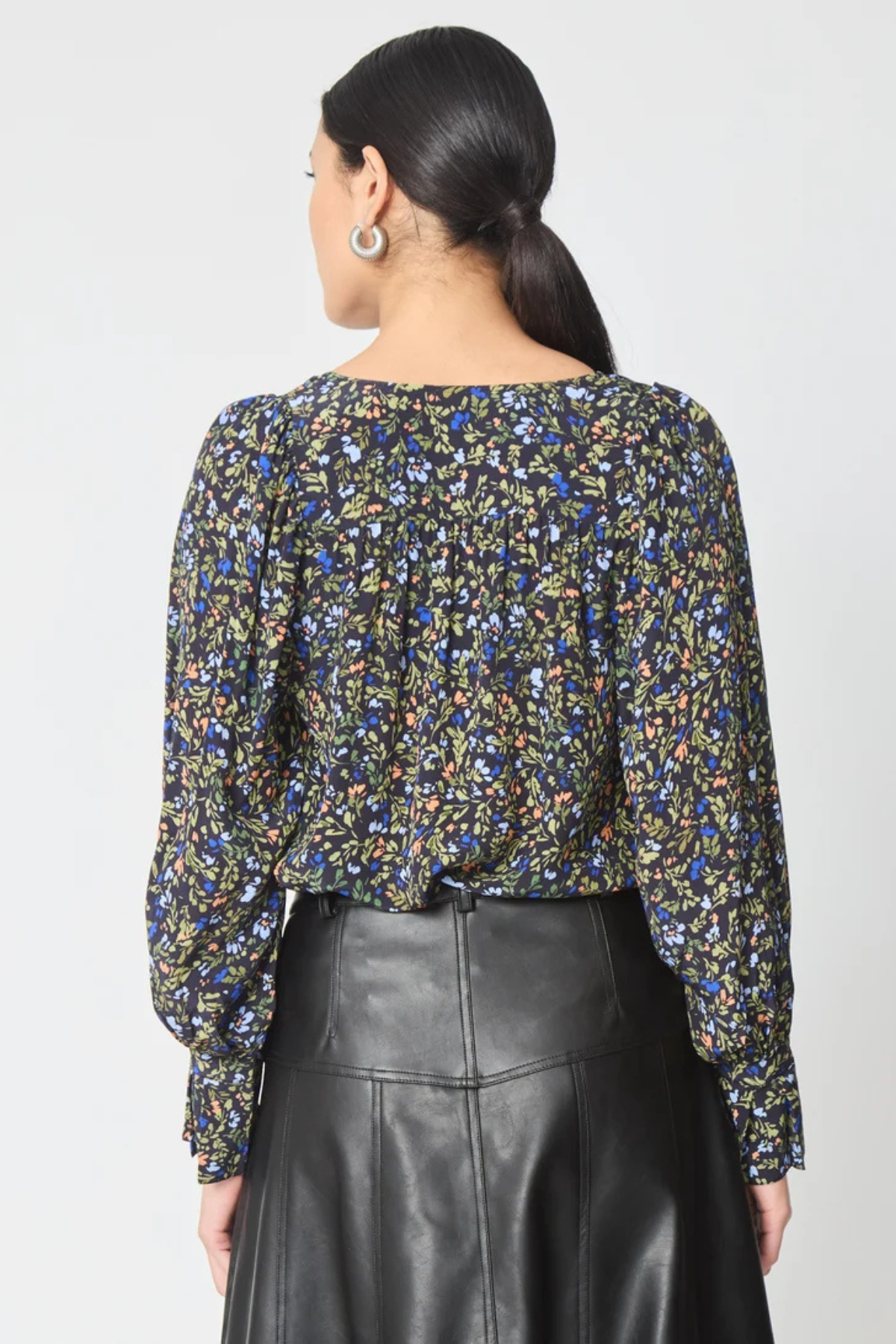 A woman shown from the back wearing a long-sleeve blouse with a dense multicolor floral print on a dark background, paired with a black leather skirt, against a light gray studio background.