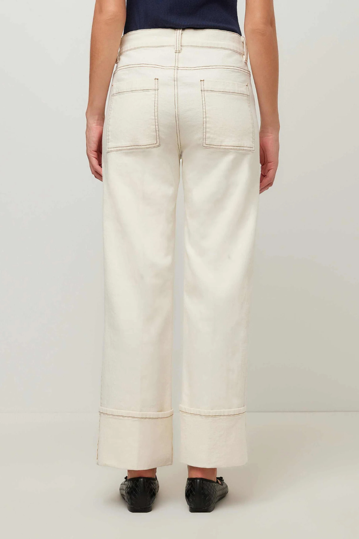 A person shown from the back wearing off-white wide-leg cropped jeans with rolled cuffs and black flats, against a plain light gray studio background.