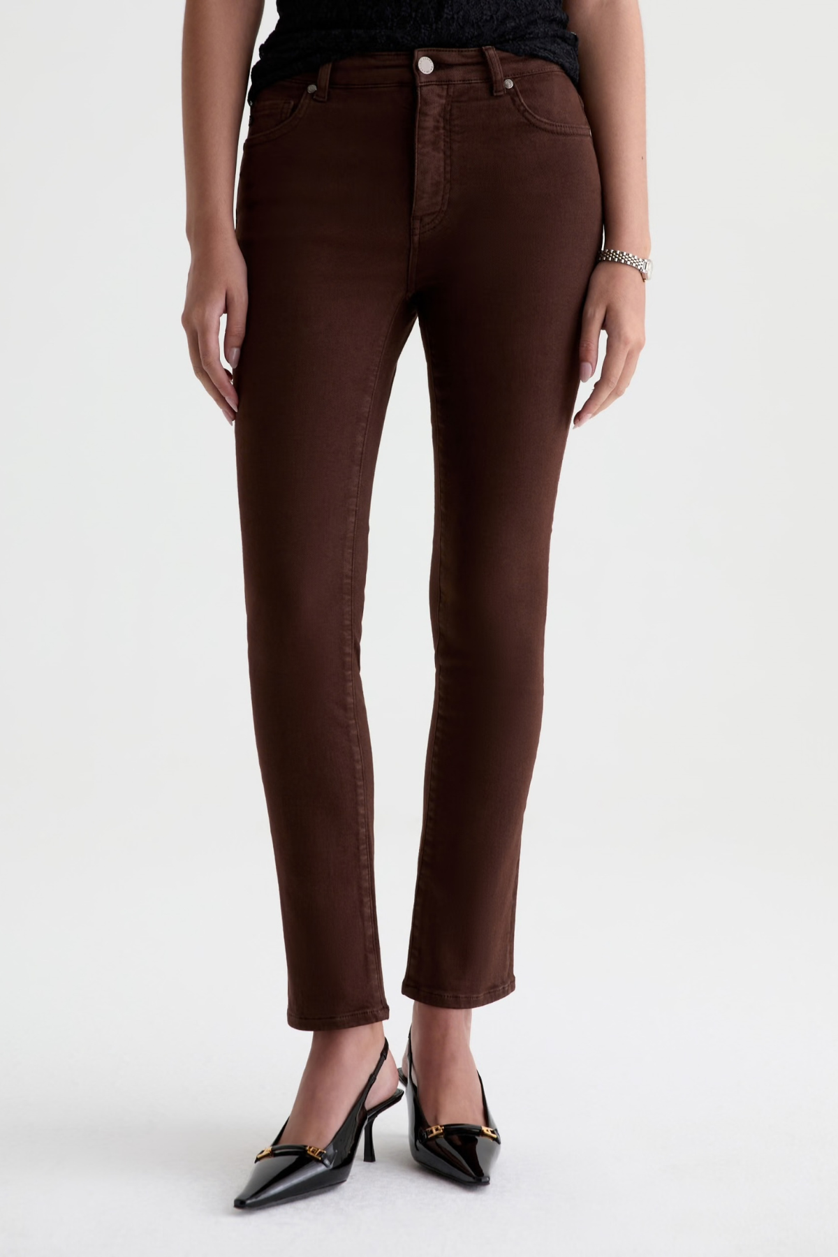 A model wearing dark brown skinny jeans and black pointed-toe heels, shown from the waist down against a plain white studio background.