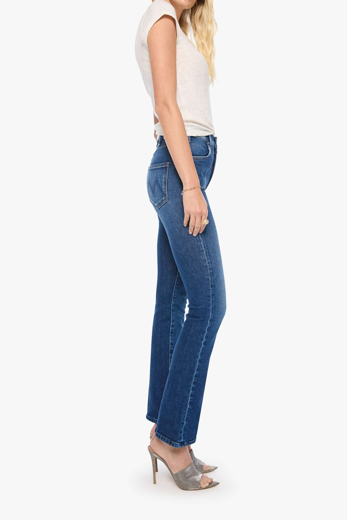 A woman shown in side profile wearing blue flare jeans, a light beige sleeveless top, and gray heeled sandals against a plain light gray studio background.