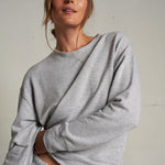Woman wearing a light grey long-sleeve knit top with a relaxed fit, photographed against a soft neutral backdrop.