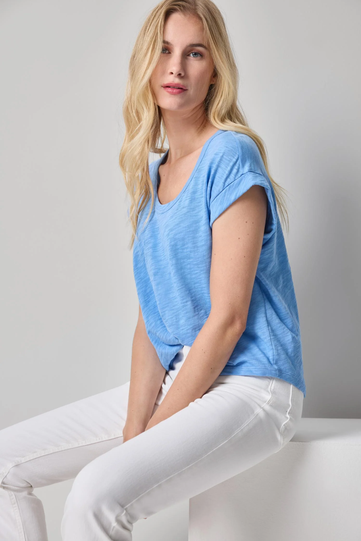 Woman wearing a blue t-shirt and white pants sitting on a white surface with a gray background