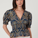 A woman modeling a navy blue short-sleeve blouse with a yellow and gold palm tree pattern, paired with white pants, against a light gray studio background.