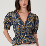 A woman modeling a navy blue short-sleeve blouse with a yellow and gold palm tree pattern, paired with white pants, against a light gray studio background.