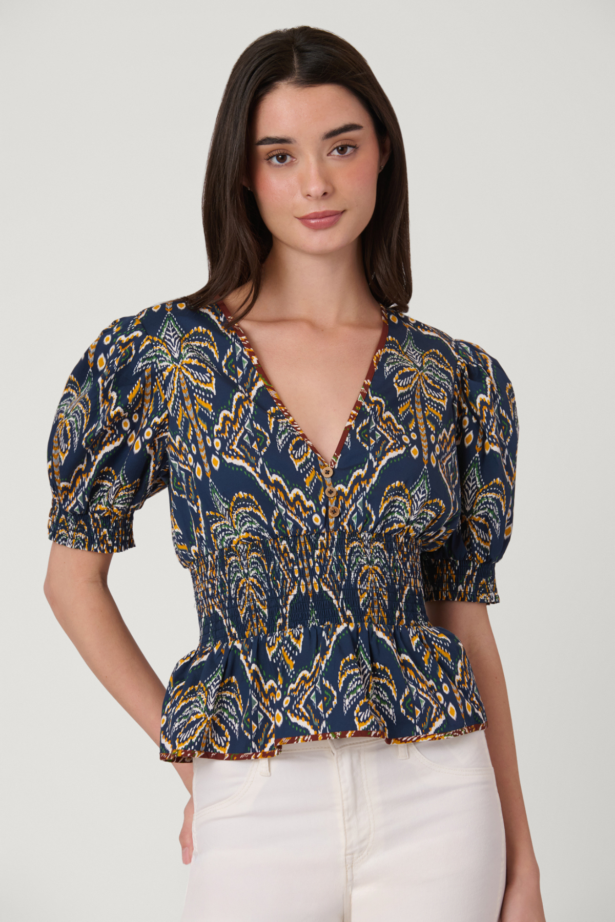 A woman modeling a navy blue short-sleeve blouse with a yellow and gold palm tree pattern, paired with white pants, against a light gray studio background.