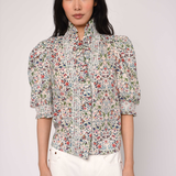 Woman wearing a floral blouse and white pants against a plain background