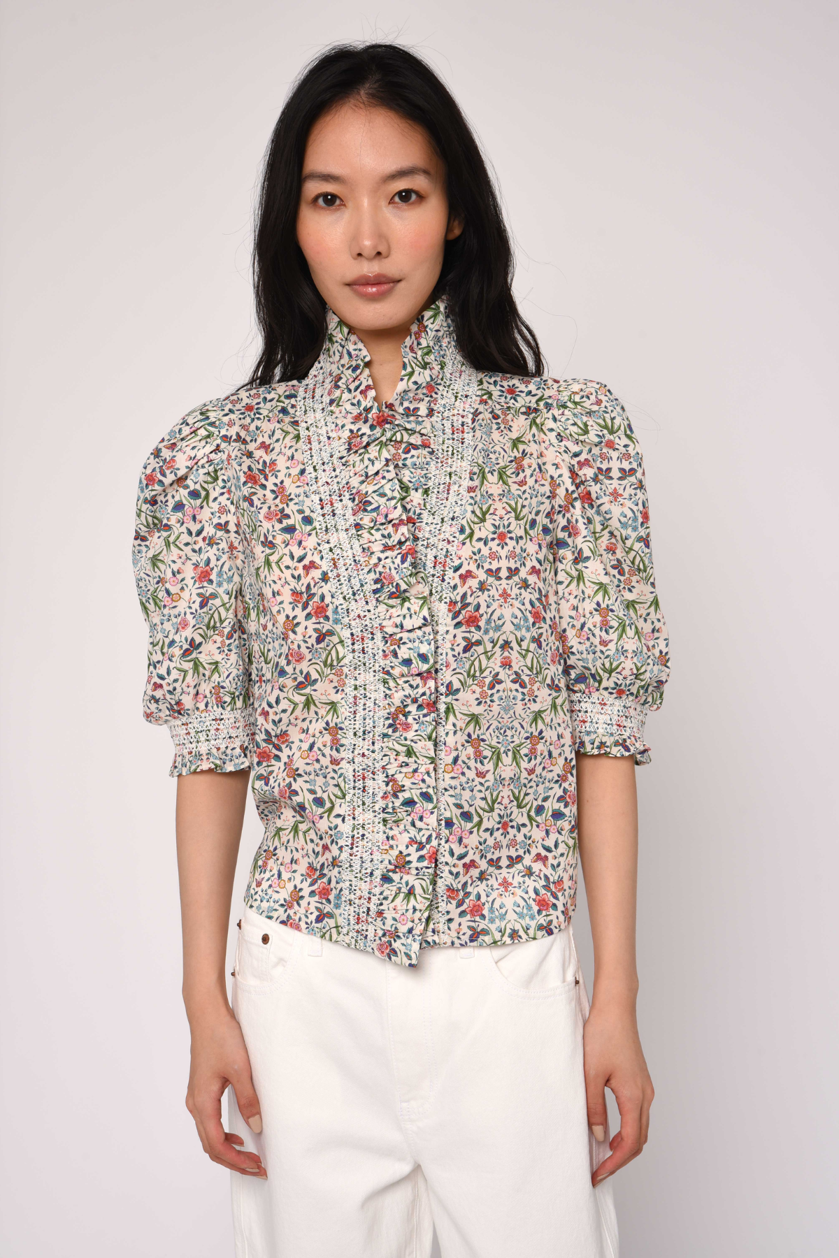 Woman wearing a floral blouse and white pants against a plain background