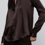 A close-up of a woman wearing a dark brown satin button-up shirt and dark brown pants against a light gray studio background.