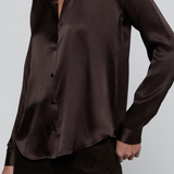 A close-up of a woman wearing a dark brown satin button-up shirt and dark brown pants against a light gray studio background.
