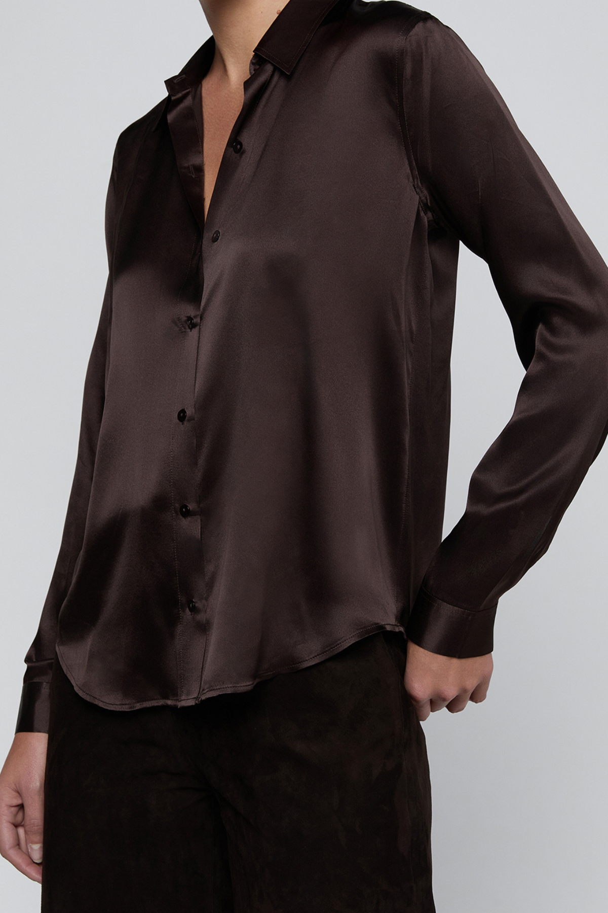 A close-up of a woman wearing a dark brown satin button-up shirt and dark brown pants against a light gray studio background.