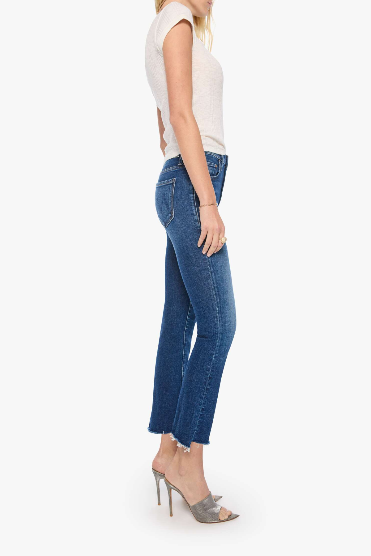 A side-profile view of a model wearing blue cropped ankle jeans, a light beige sleeveless top, and gray high-heeled sandals against a plain white studio background.