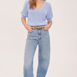 A woman modeling a light blue ribbed knit short-sleeve V-neck top paired with light-wash wide-leg jeans and a tan belt, standing against a plain light beige studio background.